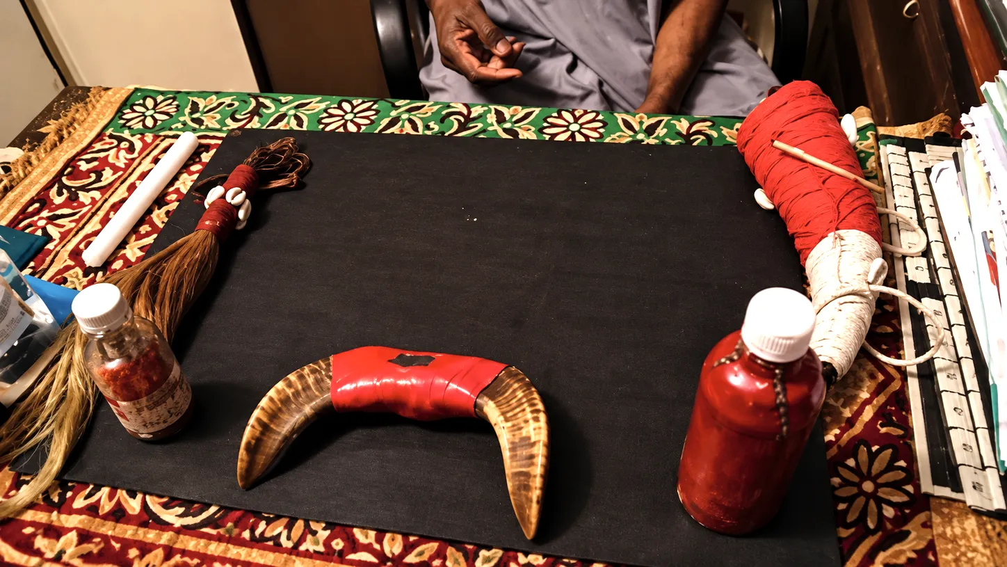 Table de consultation de Marabout Kanfory : sur un plan noir posé sur un tapis fleuri rouge, vert et ocre, une corne enrubannée de rouge, un chasse-mouches à cauris, une bougie blanche, des flacons et un objet rituel noué de fil rouge, le praticien assis derrière